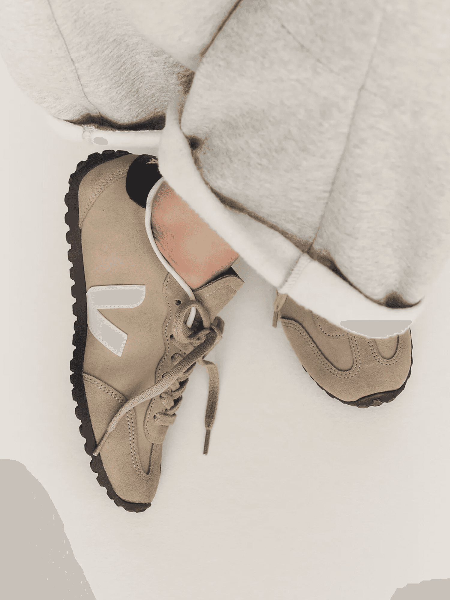 Beige sneakers with white logo, black soles, and gray pants cropped in white background.