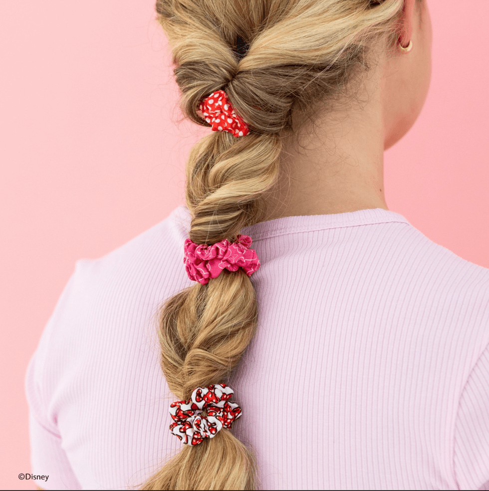 Blonde hair in a braid with red and pink scrunchies against a pink background.