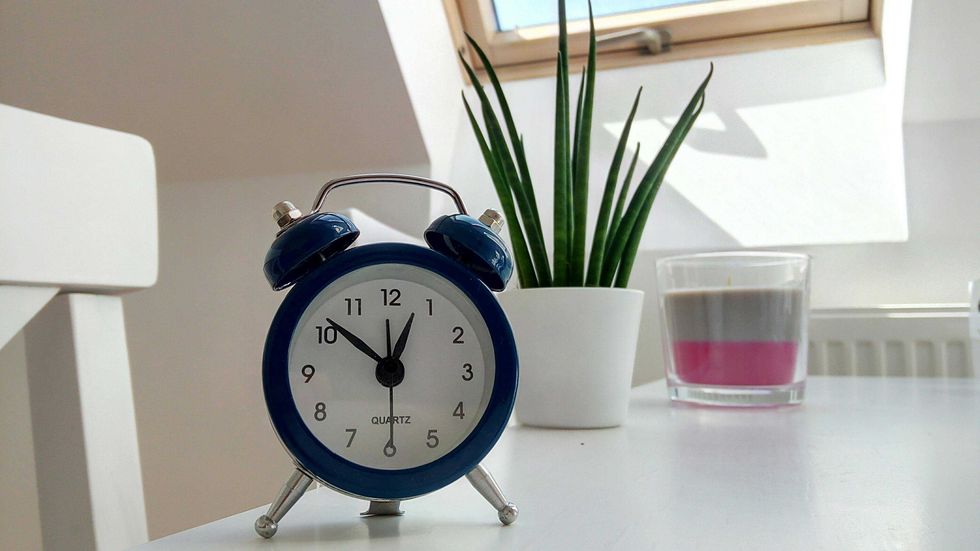 Blue alarm clock, plant, and candle on a white table by a sunny window.