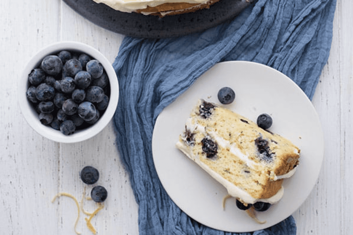 Blueberry cake with a slice on a plate, garnished with blueberries and lemon zest.