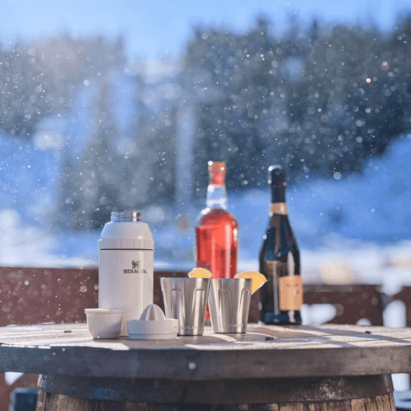 Bottles and cups on snowy barrel table outdoors in sunlight.