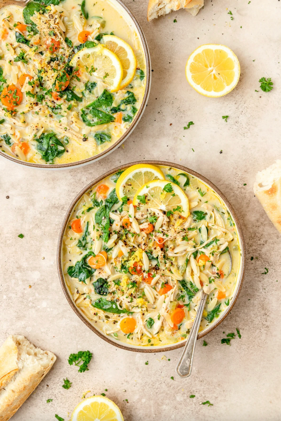 Bowl of creamy soup with lemon slices, carrots, spinach, and herbs on a beige surface.