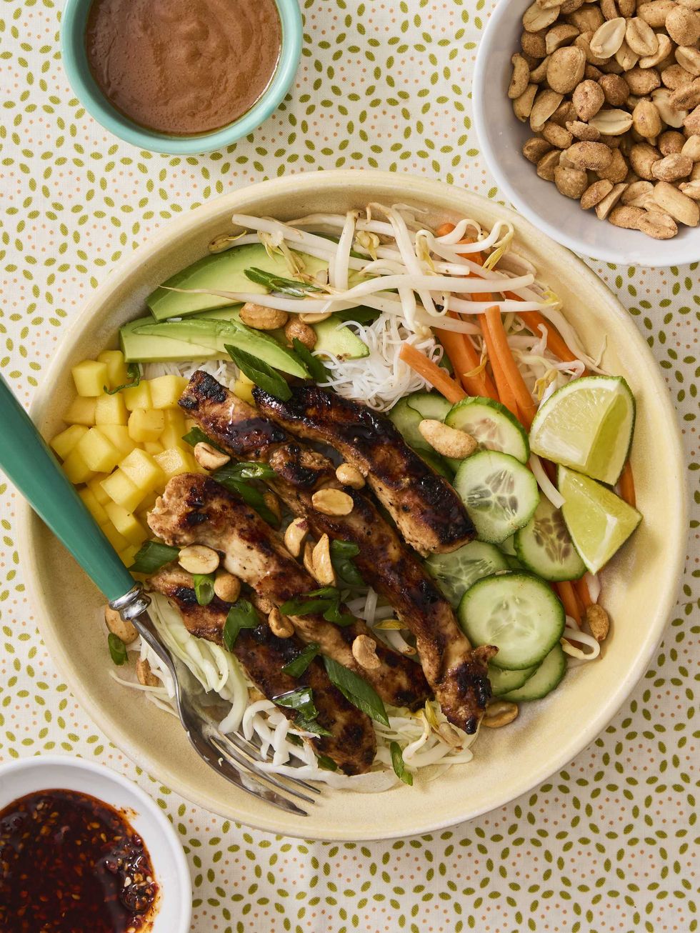Bowl of grilled chicken, veggies, noodles, mango, and peanuts with sauces on a patterned tablecloth.