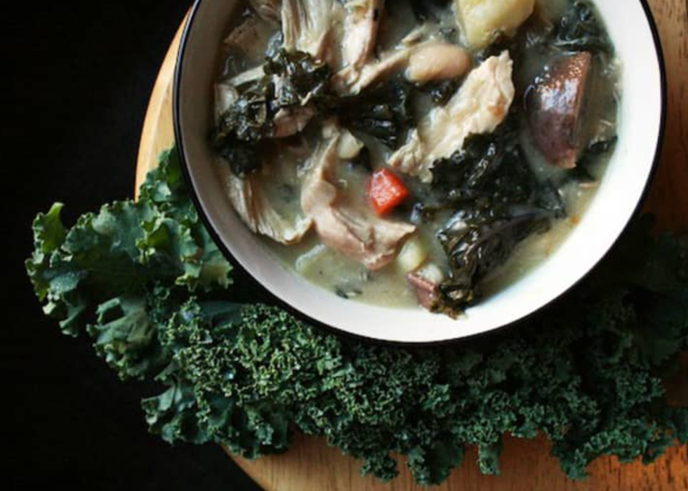 Bowl of hearty stew with chicken, kale, carrots, and beans on a wooden board with fresh kale.