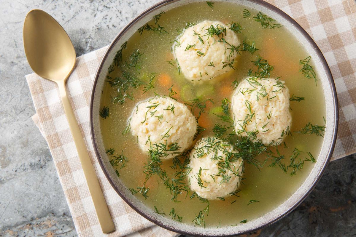 Bowl of matzo ball soup with fresh dill and a gold spoon on checkered cloth.