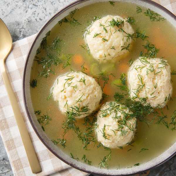 Bowl of matzo ball soup with fresh dill and a gold spoon on checkered cloth.