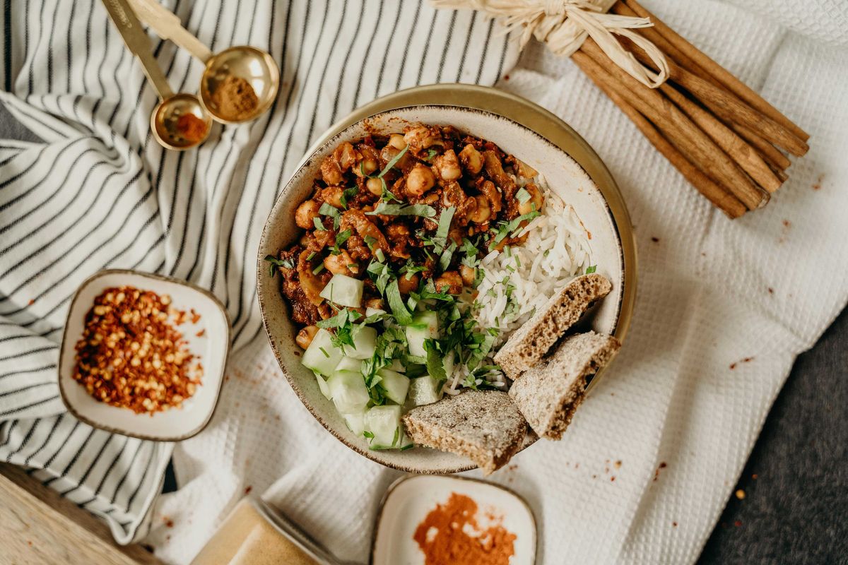 Bowl of rice, chickpeas, cucumber, herbs, and bread, with spices on a striped cloth.
