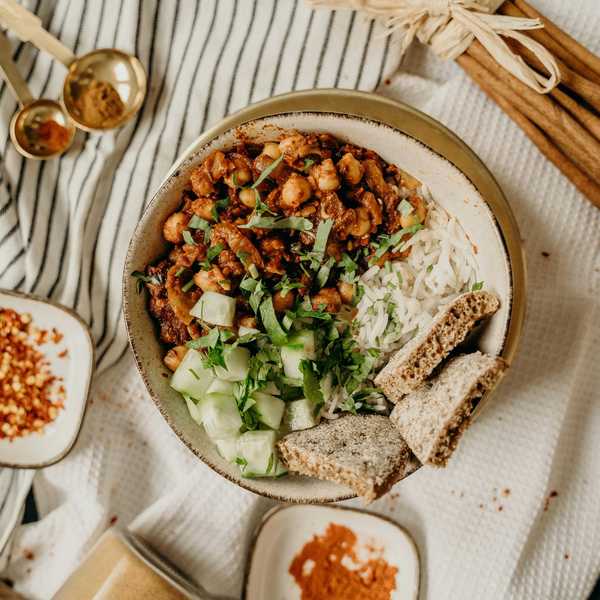 Bowl of rice, chickpeas, cucumber, herbs, and bread, with spices on a striped cloth.