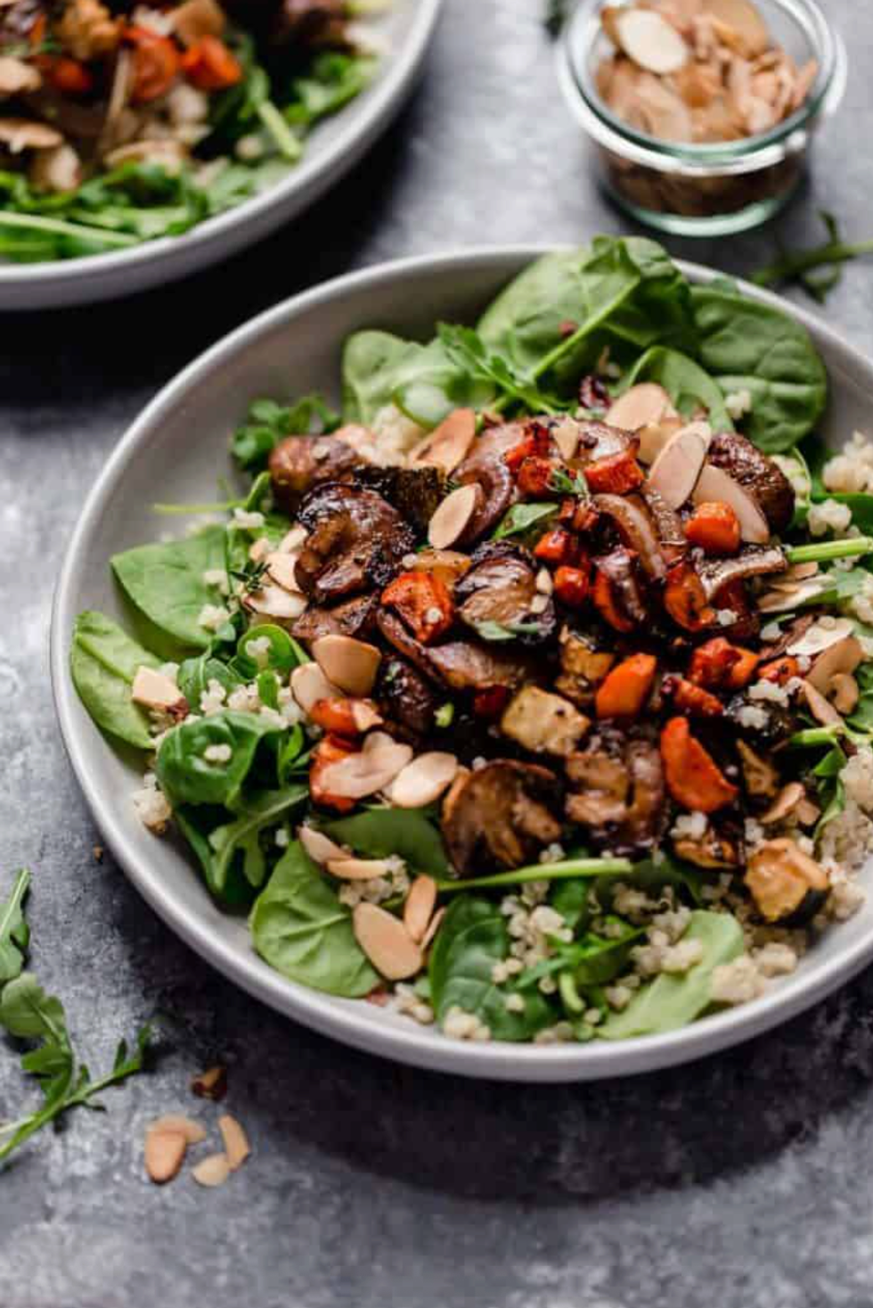 Bowl of roasted vegetables, greens, quinoa, and almonds on a gray surface.