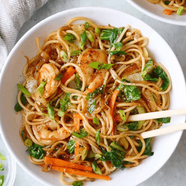 Bowl of shrimp lo mein with vegetables and chopsticks on a light surface.