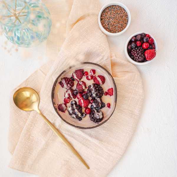 Bowl of smoothie with berries on cloth, next to seeds and berries, with a gold spoon.