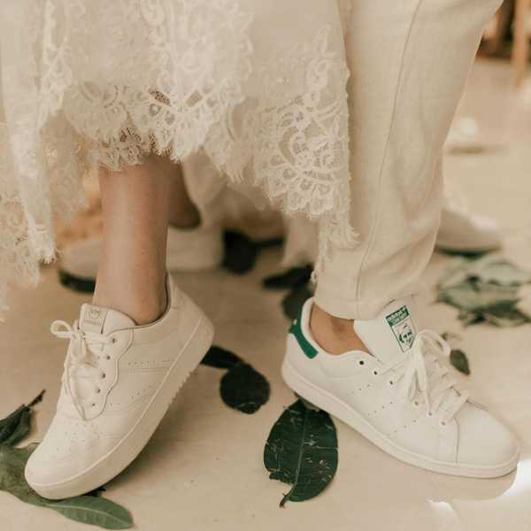 Bride and groom in white sneakers standing on scattered green leaves.
