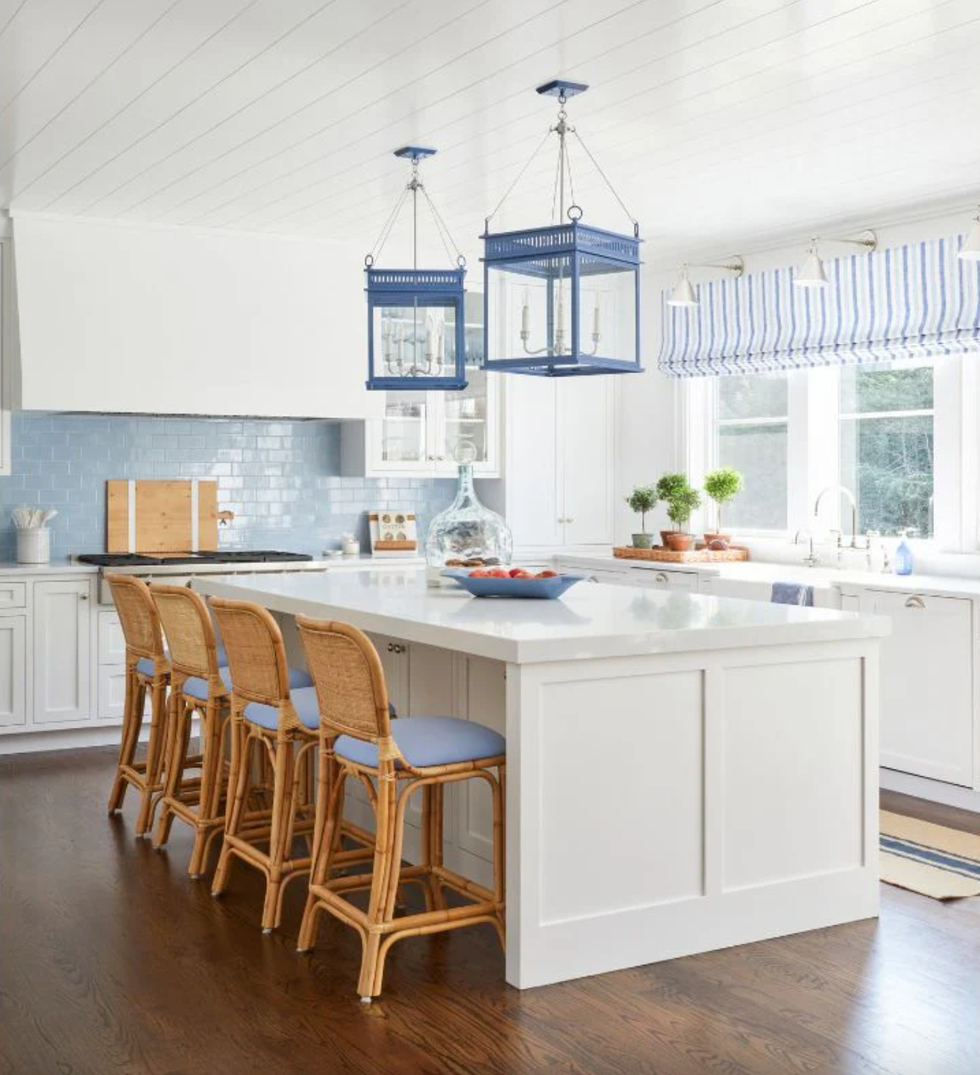 Bright kitchen with white island, wicker stools, blue accents, and large windows.