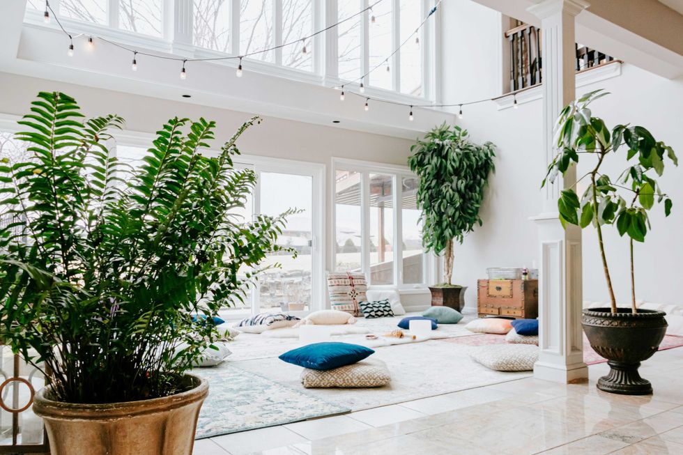 Bright room with large plants, cushions on the floor, string lights, and tall windows.