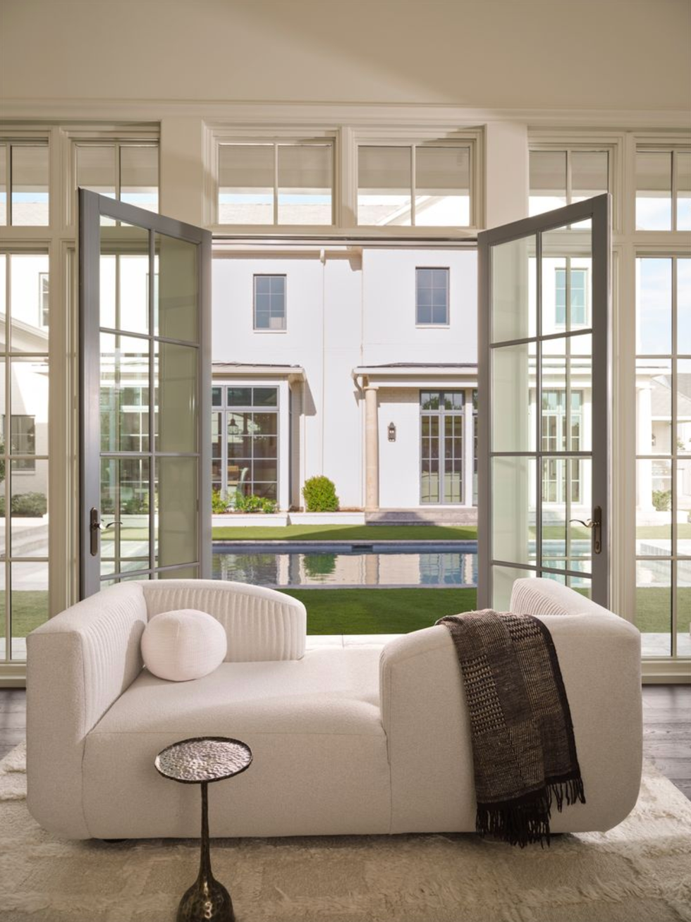 Bright room with white sofa, open doors, and view of pool and modern house outside.