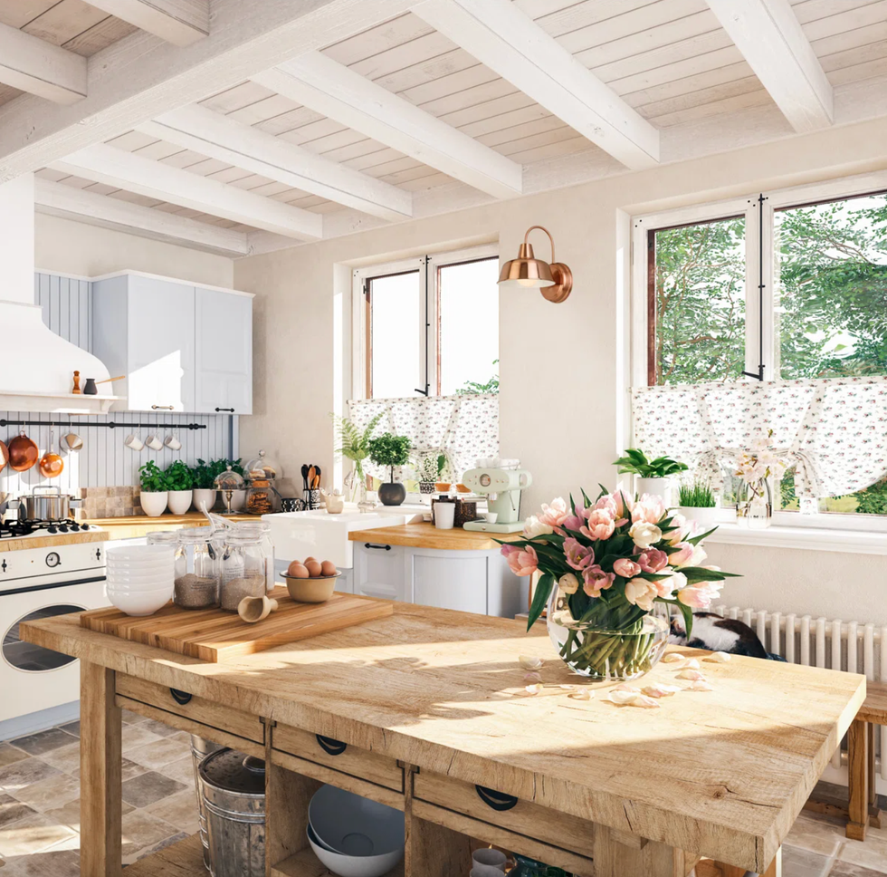 Bright rustic kitchen with wooden island, flowers, and natural light from large windows.