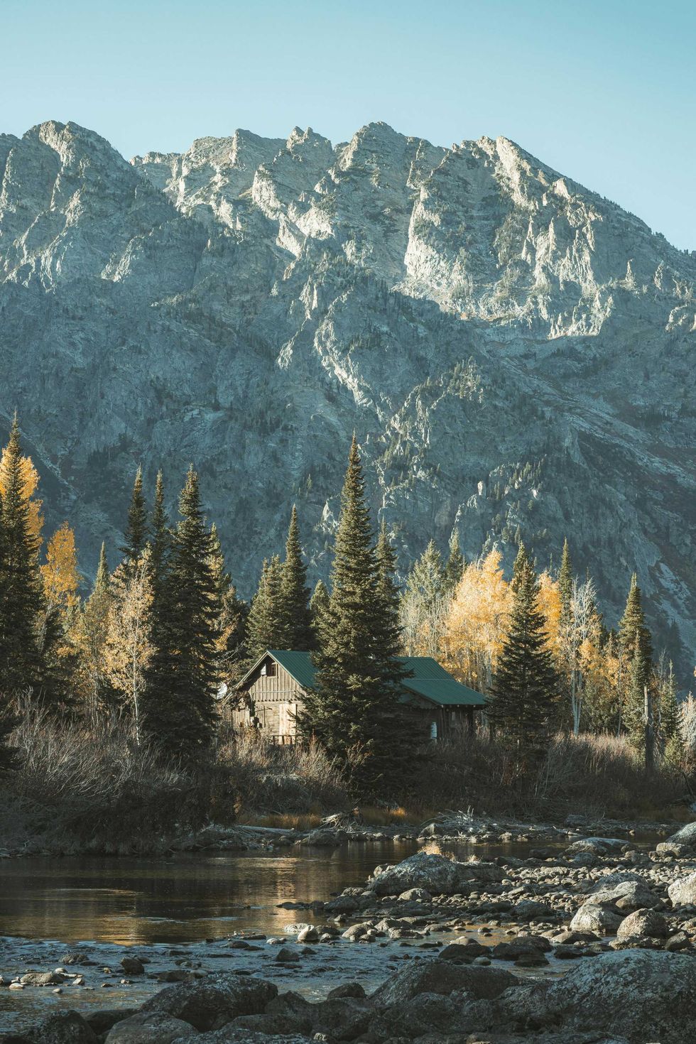Cabin nestled in forest, with river and mountains in the background.