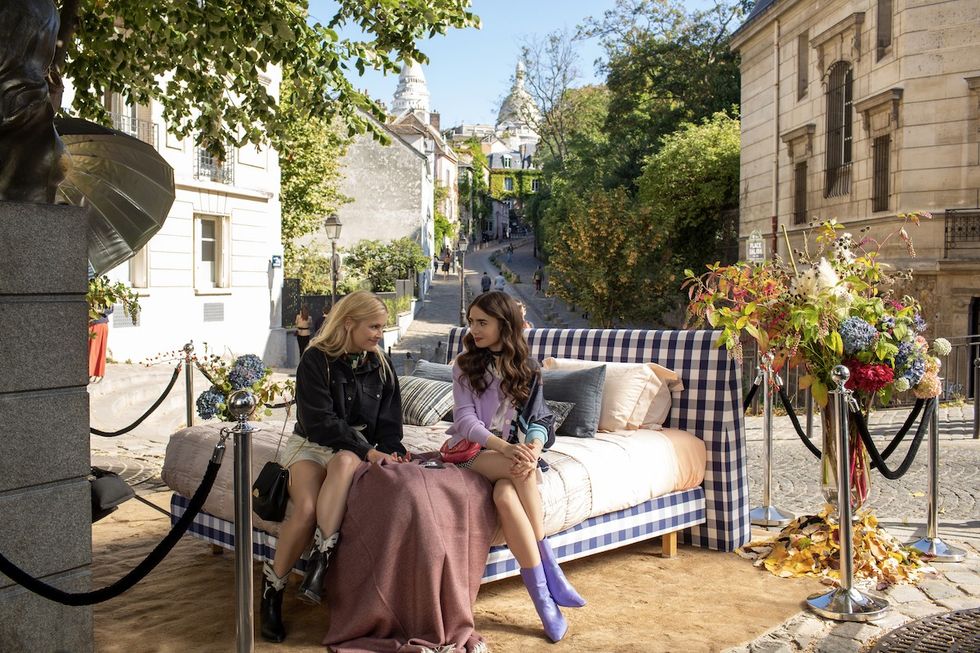 Camille Razat as Camille and Lily Collins as Emily sitting on a bed in paris
