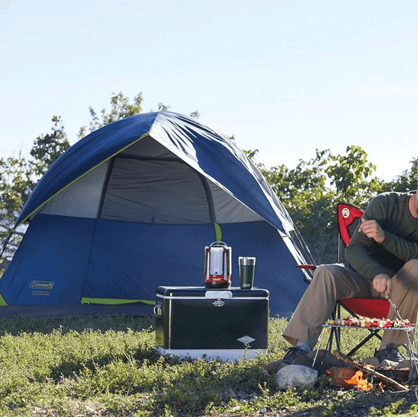 Camping scene with a blue tent, cooler, and person roasting food over a campfire.