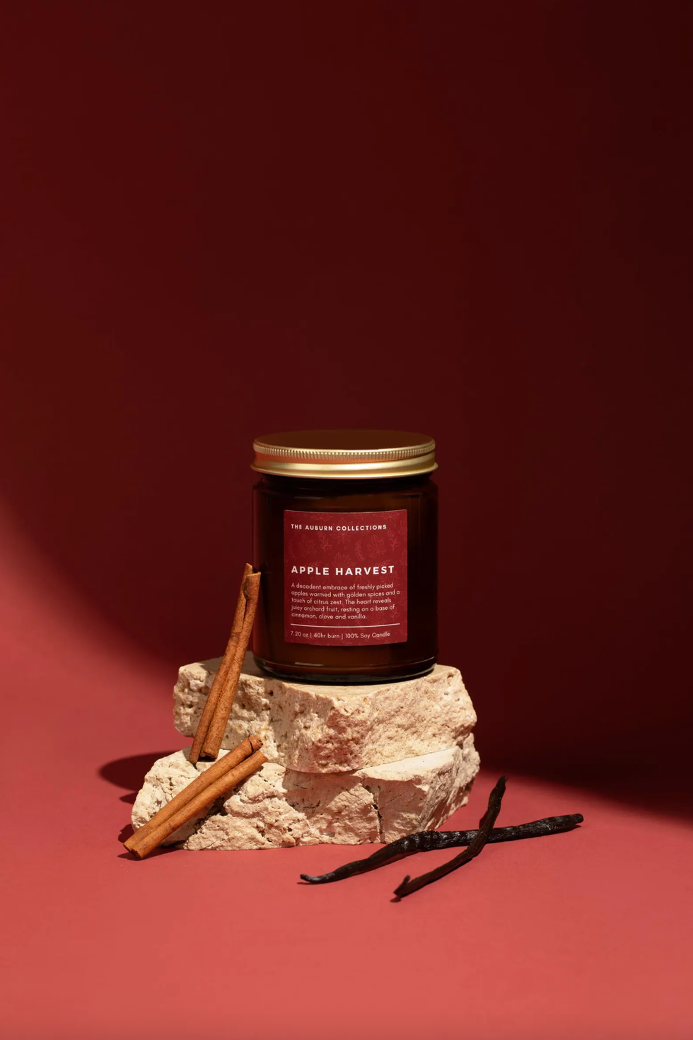 Candle labeled "Apple Harvest" with cinnamon sticks and vanilla on stone against red background.