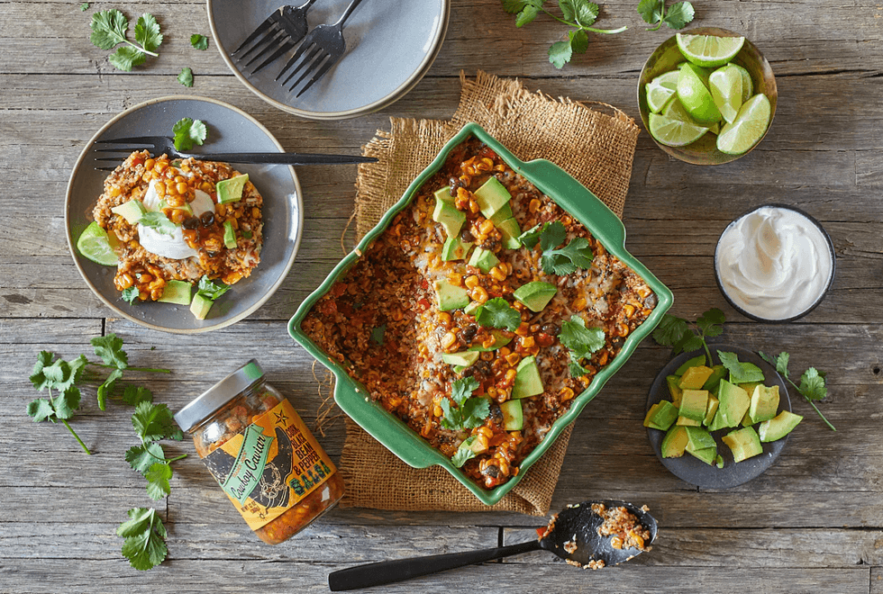 Casserole with avocado and corn in dish, plate with slice, lime wedges, and sour cream on table.