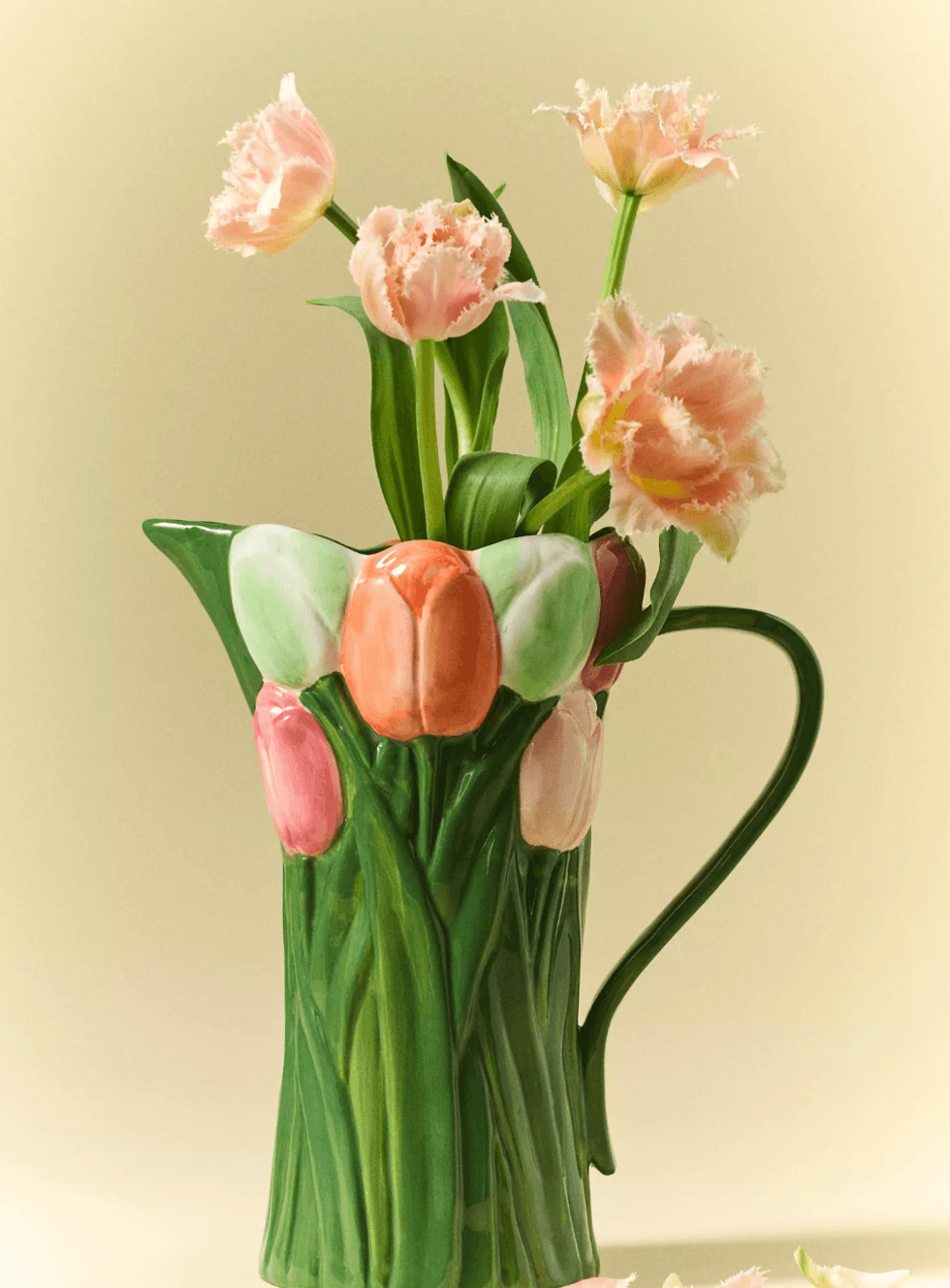 Ceramic pitcher with tulip designs, holding pink frilled tulips against a beige background.