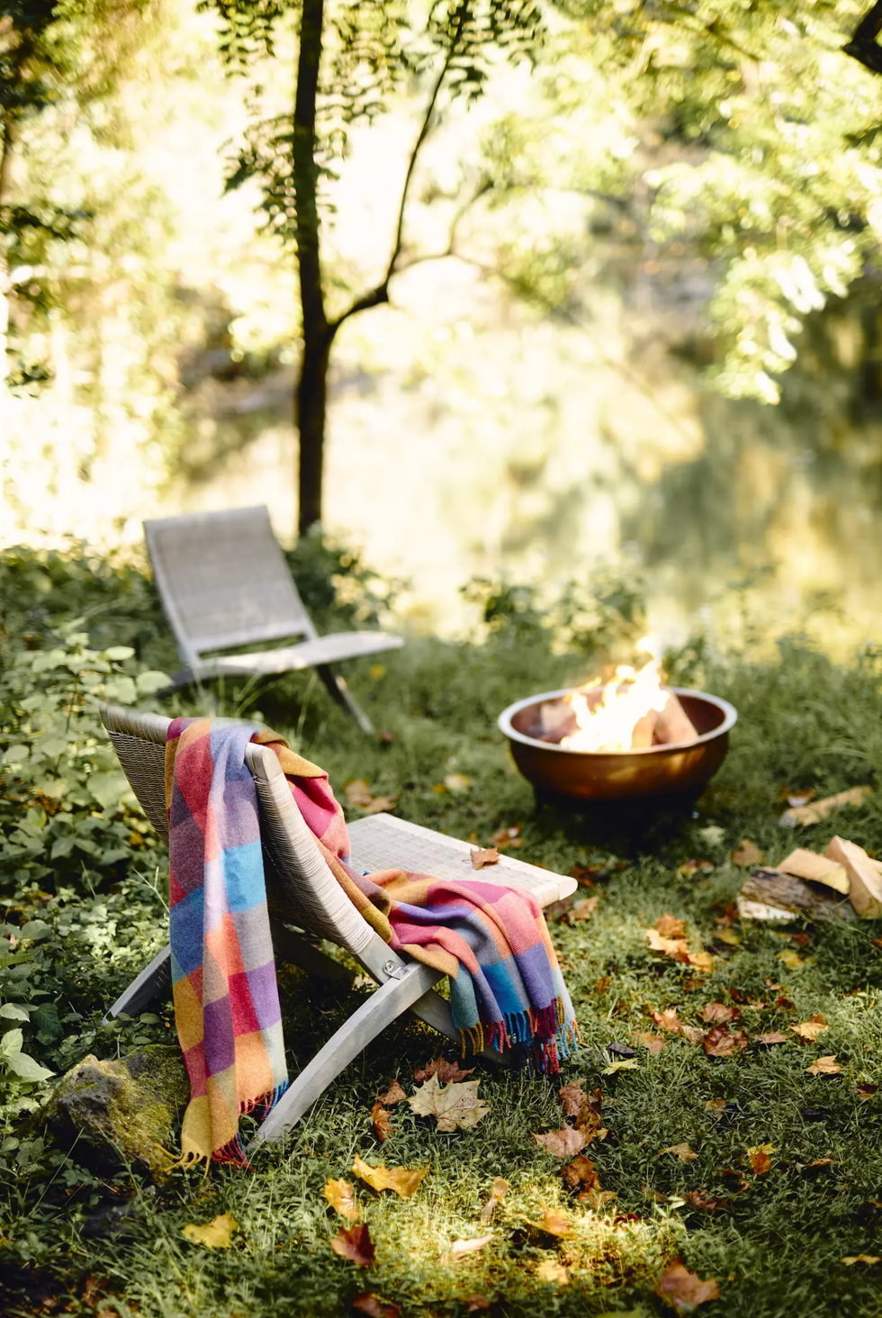 Chairs with a colorful blanket by a fire pit in a lush, leafy garden setting.