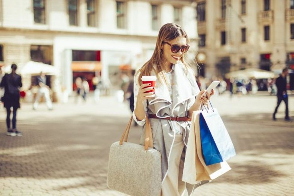 Cheerful woman is shopping in the city. She is holding a coffee to go and using her smart phone.