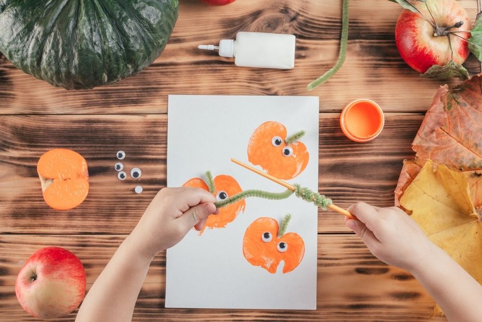 Child crafting pumpkin art with paint, googly eyes, apples, and leaves on a wooden table.