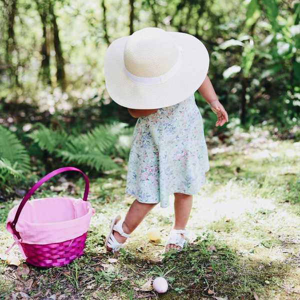 Child in a floral dress and hat, standing next to a pink basket in a sunny forest.