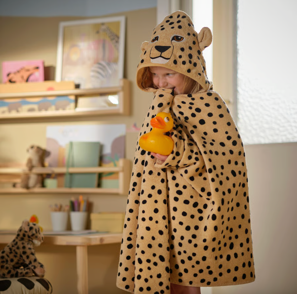 Child in a spotted hooded towel, holding a rubber duck, smiles playfully indoors.