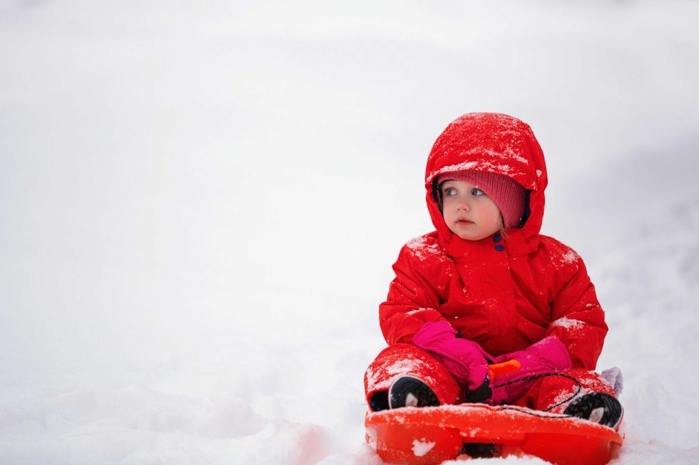 Child in red snowsuit sitting on a sled in the snow, wearing pink mittens.