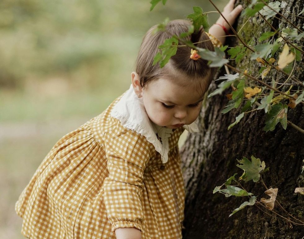 Child in yellow dress exploring beside a tree in a forest landscape.