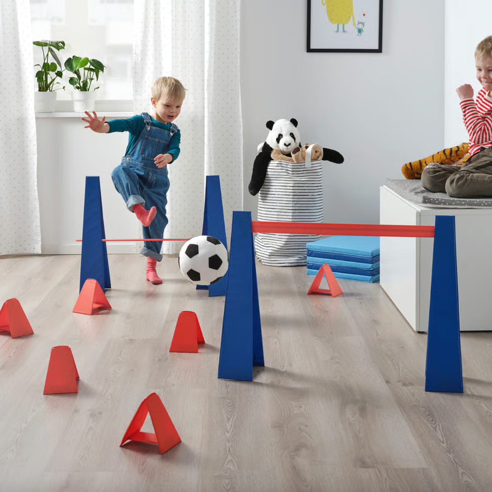 Child playing soccer indoors with an obstacle course; another child watches nearby.