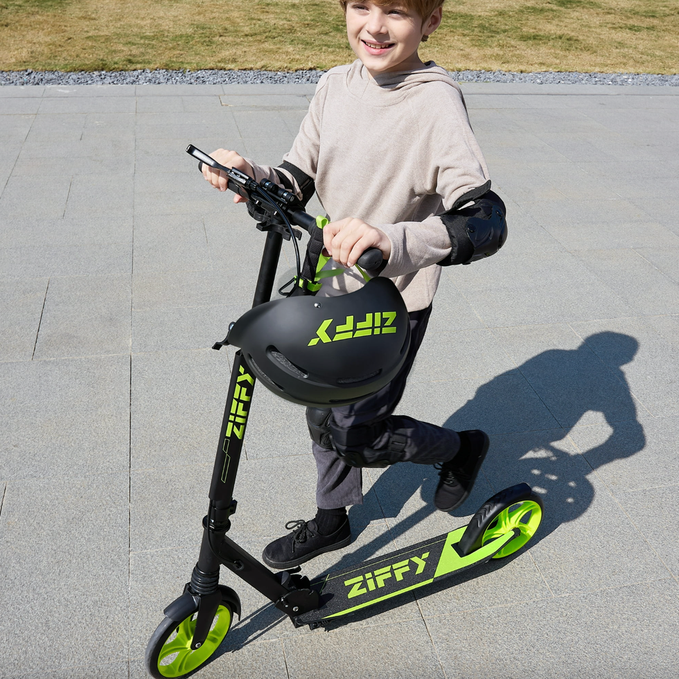 Child riding a black and green scooter, holding a matching helmet, smiling in sunlight.
