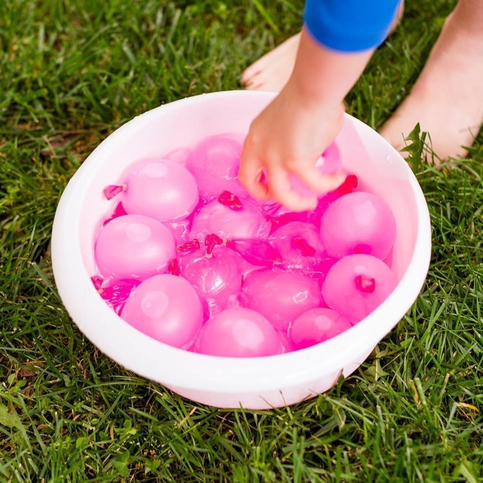 Child's hand grabbing pink water balloon from bucket on grass.