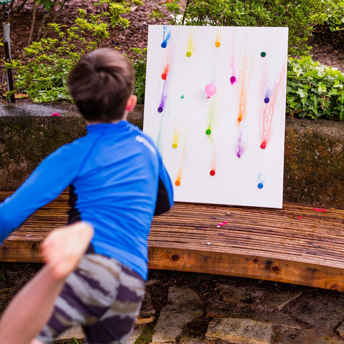 Child throwing at paint-filled balloons on canvas on a bench.