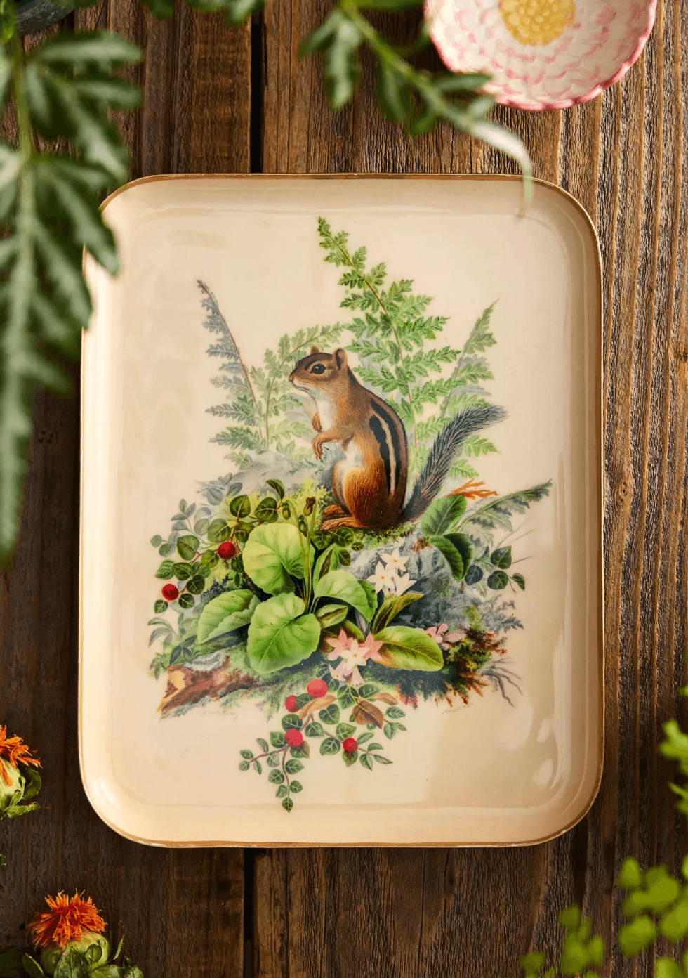 Chipmunk illustration on a tray with greenery and flowers, surrounded by wooden table decor.