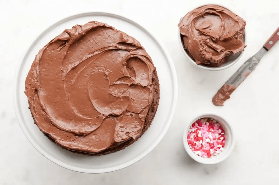 Chocolate cake with frosting, a knife, and heart-shaped sprinkles on a white surface.