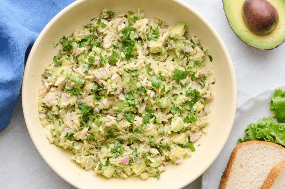 Chopped salad with avocado and herbs in a bowl, surrounded by bread and a halved avocado.