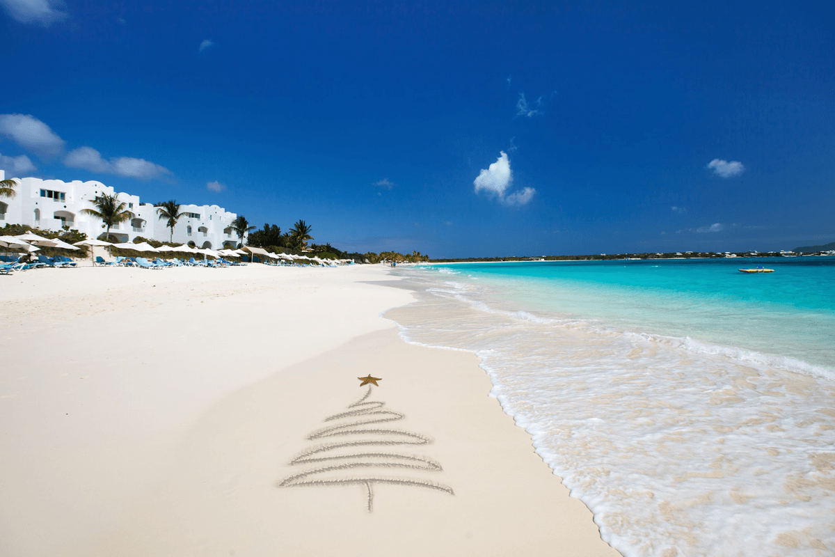 Christmas tree drawn in sand on a sunny beach, turquoise sea, white buildings in the background.