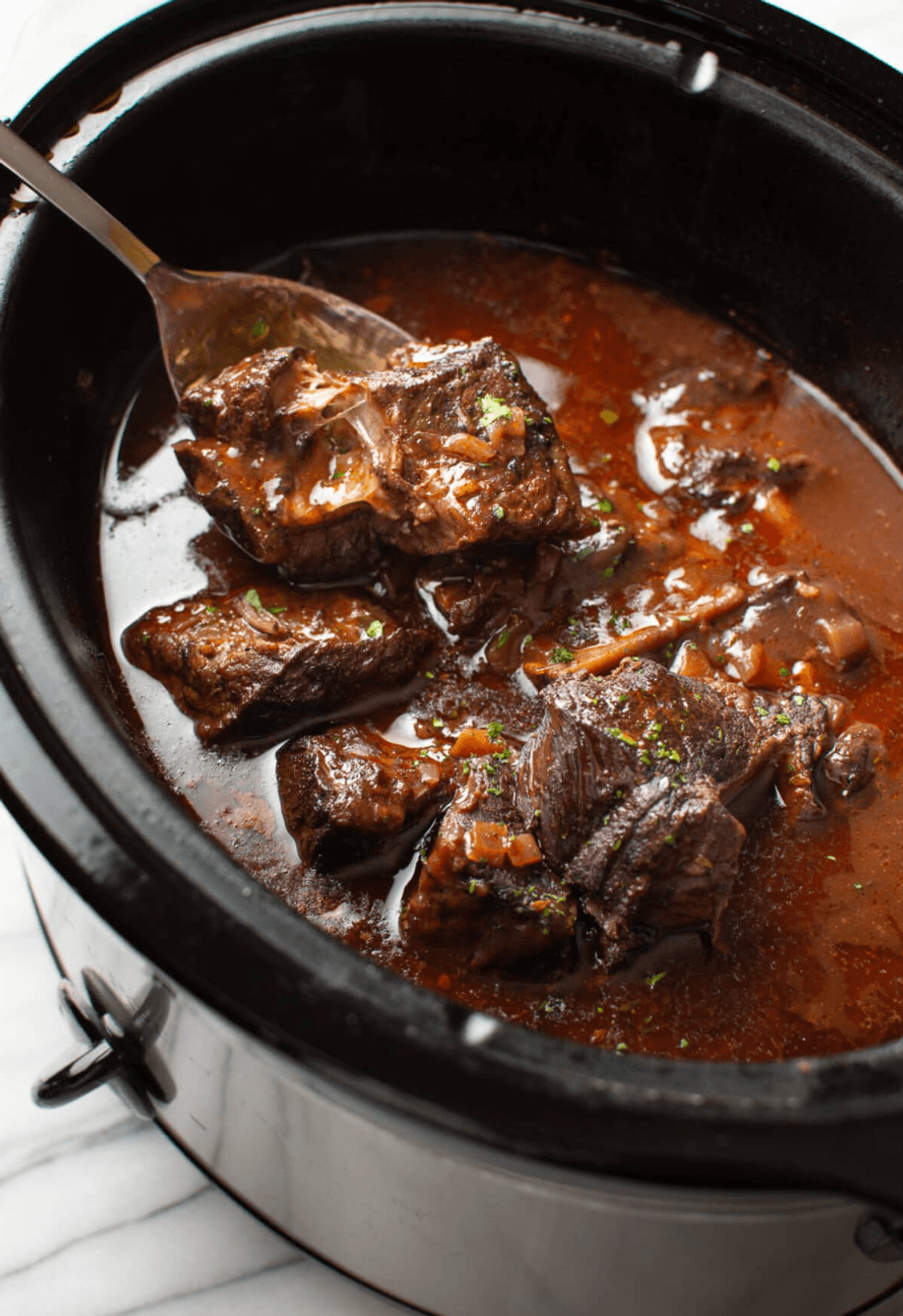 Chunks of beef in rich sauce in a slow cooker with a serving spoon.
