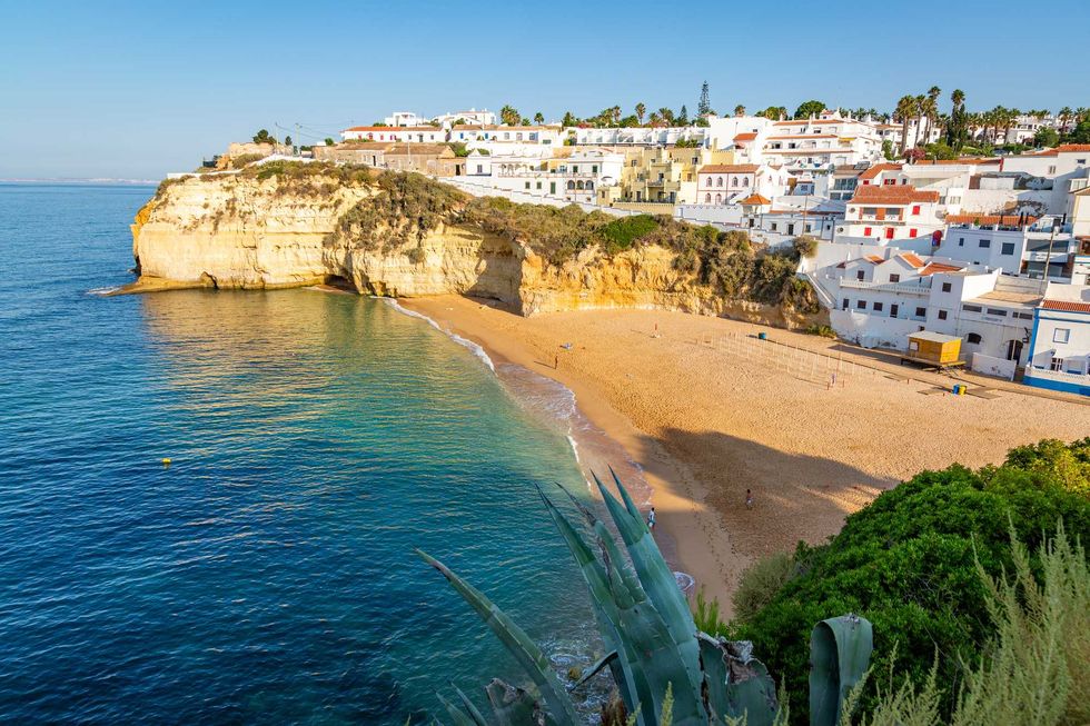 Clifftop village overlooking a sandy beach and clear blue sea under a sunny sky.