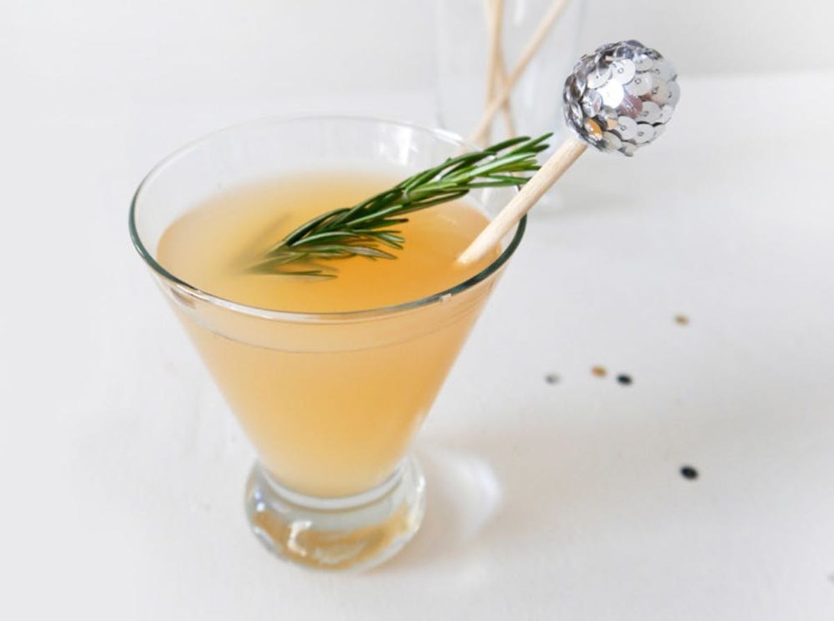 Cocktail in a martini glass with rosemary and a sparkly stir stick on a white surface.