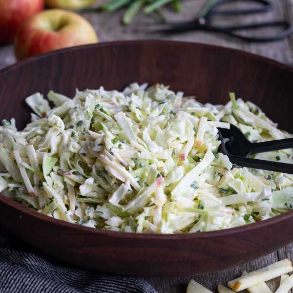 Coleslaw with apples in a wooden bowl, apples and chives in the background.