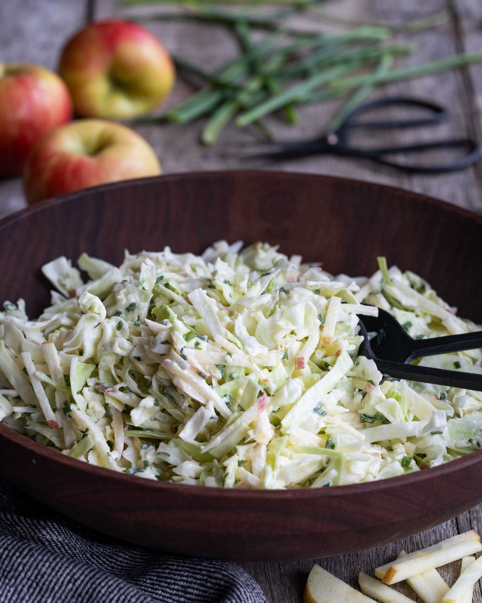 Coleslaw with apples in a wooden bowl, apples and chives in the background.