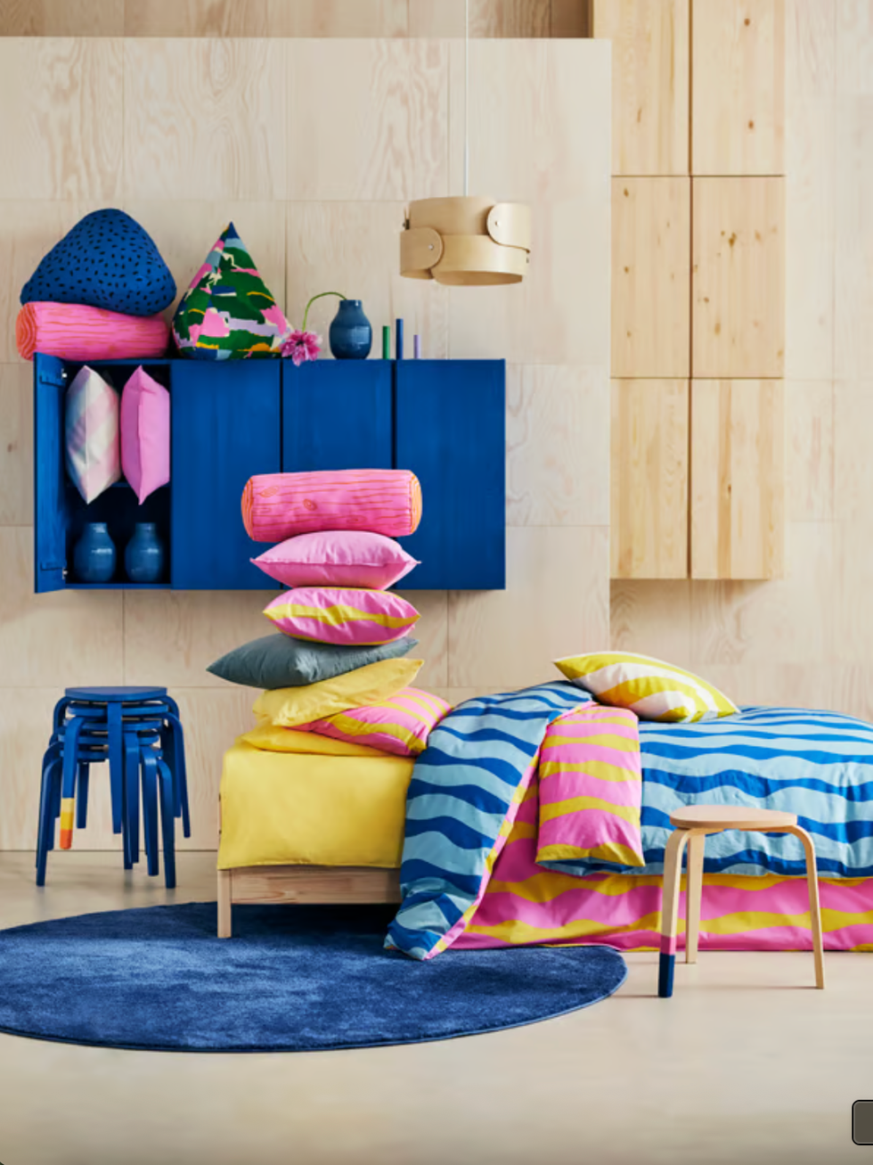 Colorful bedroom with bright bedding, pillows, blue cabinets, and stools on a circular rug.
