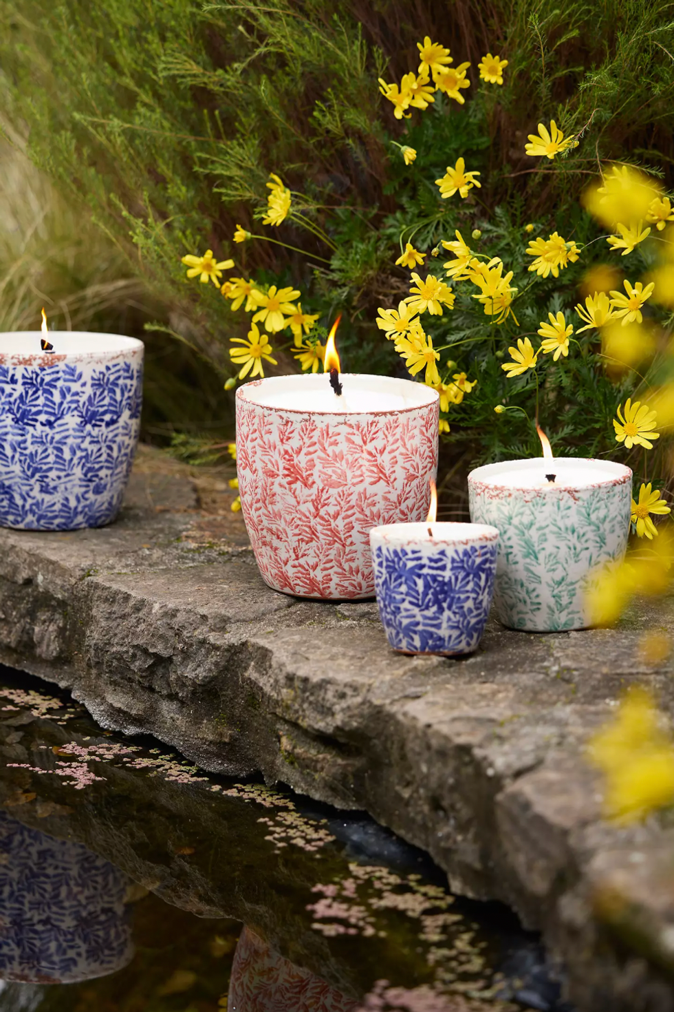 Colorful candles on stone among yellow daisies by a reflective pond.