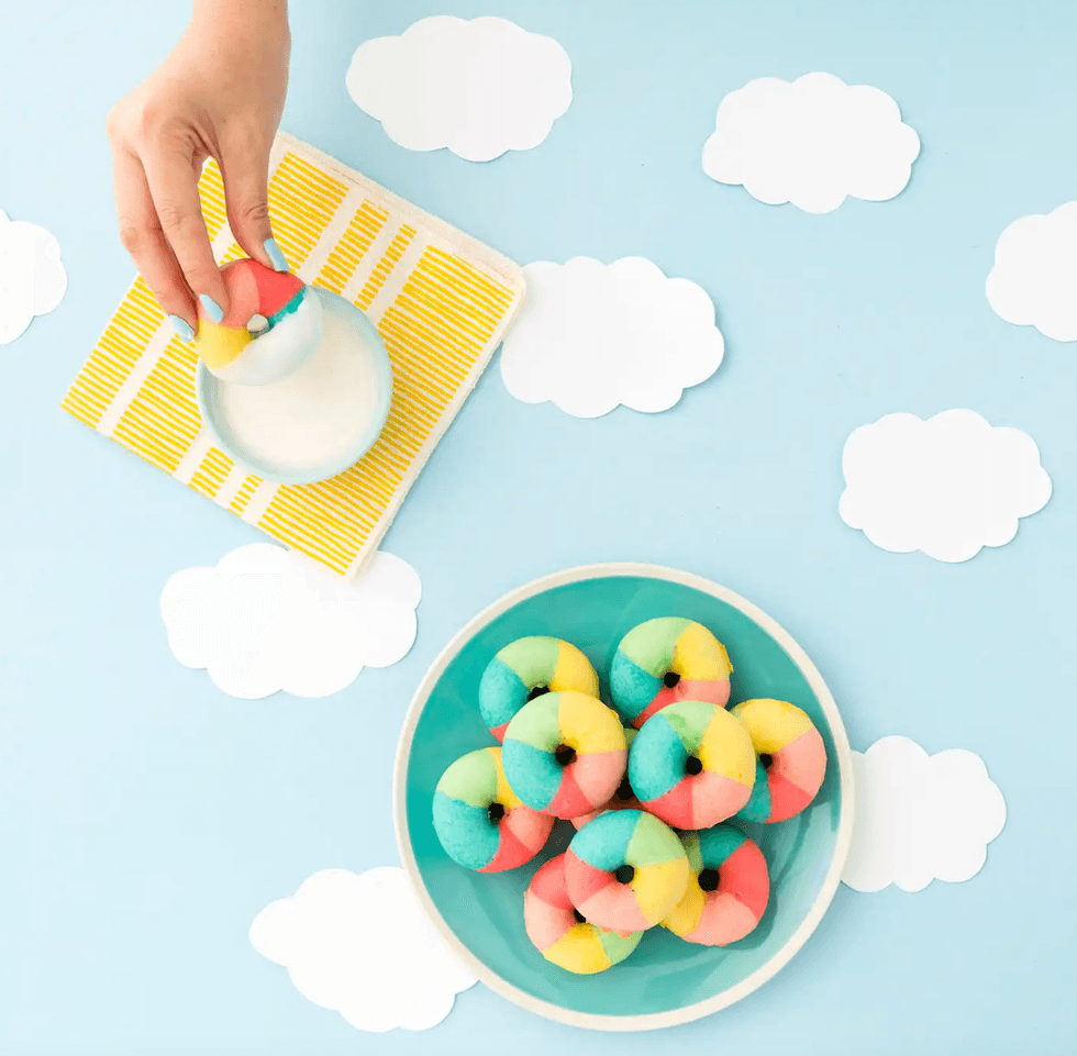 Colorful donuts on a blue plate, clouds background, one being dipped in milk.