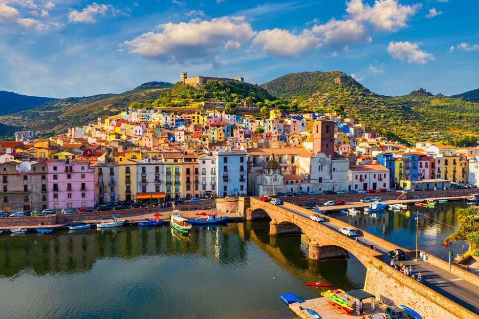 Colorful hillside town with river, arched bridge, and boats under a blue sky.